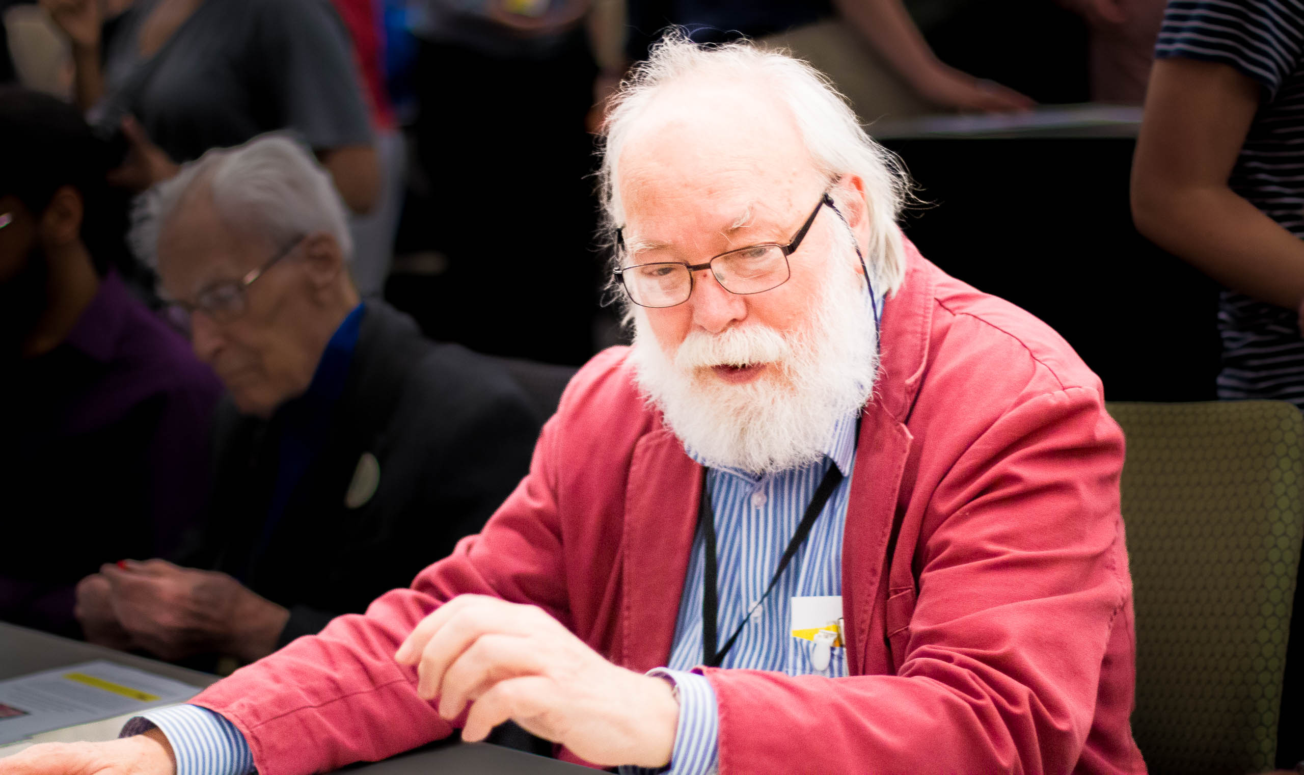 A photo of David Singmaster at a conference, looking curiously at something on a table. Richard Guy in the background on the left.